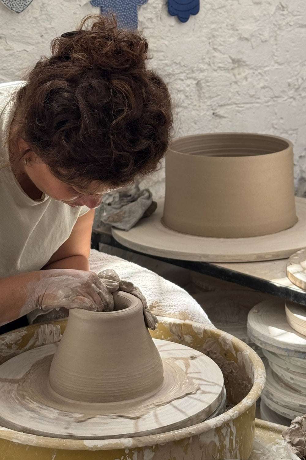 A person with curly brown hair wearing a white shirt and gloves shapes a clay pot on a pottery wheel, with another clay pot and pottery tools in the background against a white textured wall.