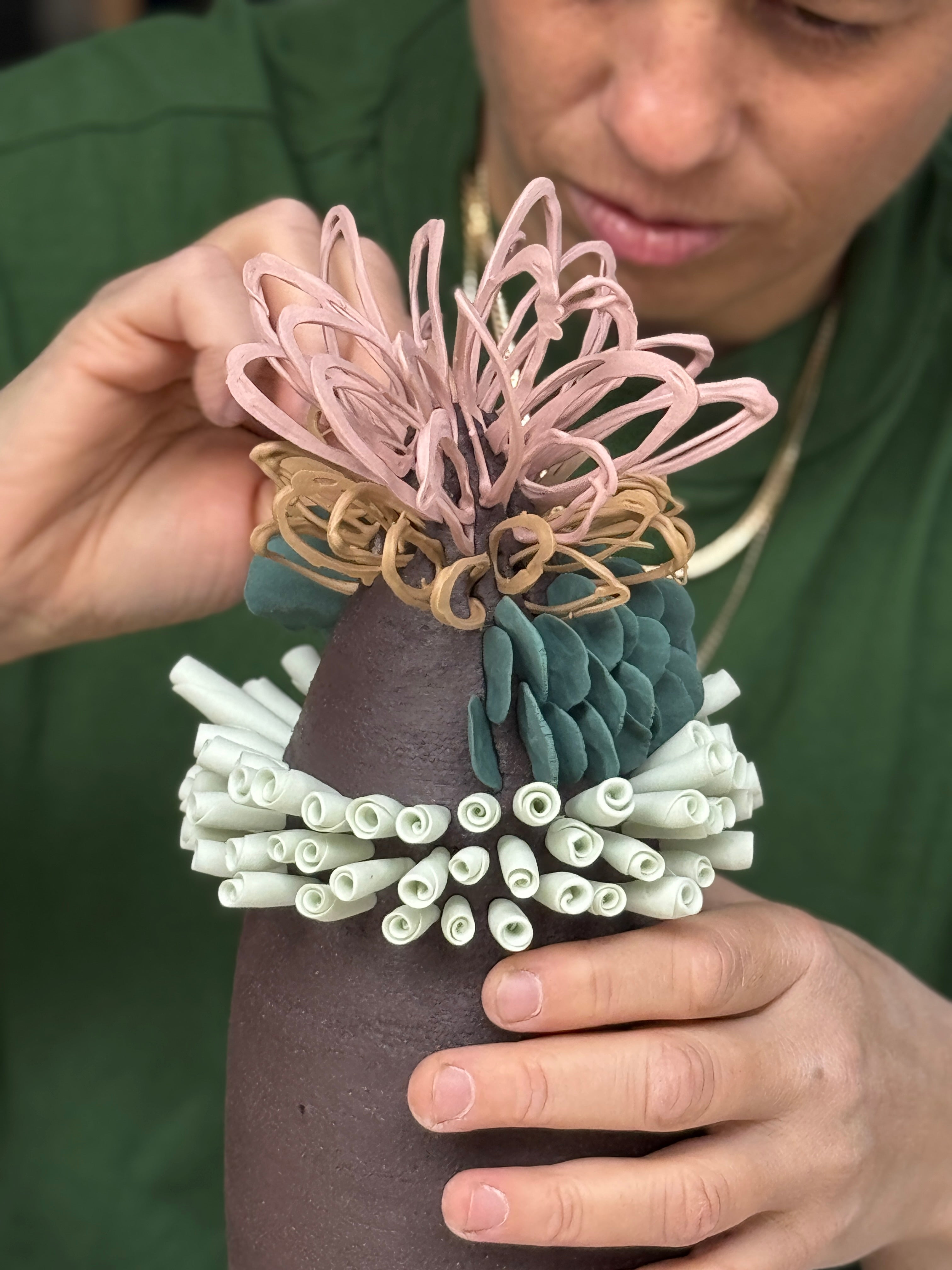 A close-up of a person holding a handmade clay pot with a textured, organic design. The pot features a dark brown body, a collar of rolled white clay, a green leaf-like section, and a pink and tan flower-like top.