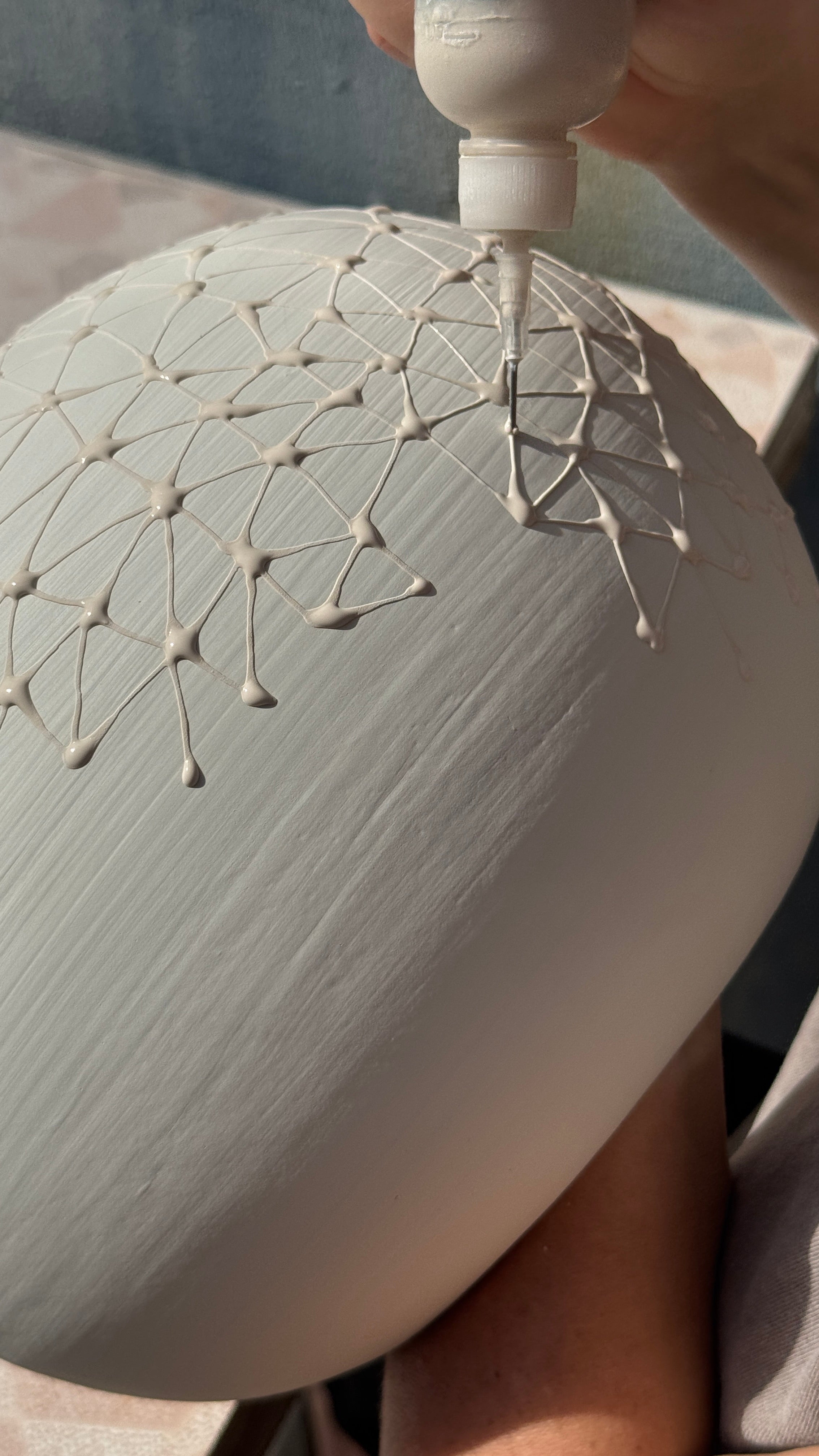 A close-up of a hand applying white decorative lines in a geometric pattern onto a large, matte, off-white ceramic or plaster sphere using a small applicator bottle.